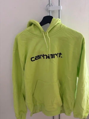 Gul/lime hoodie från Carhartt - Säljer en limegul hoodie från Carhartt med svart logga på bröstet. Hoodien har huva med dragsko och en stor magficka framtill. Tillverkad i mjuk bomull, perfekt för en chill och avslappnad stil.
