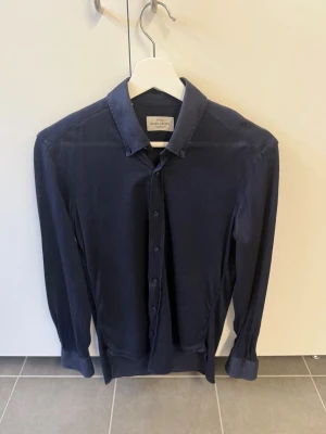 Mörkblå skjorta från Rose & Born, stl 38 - Snygg mörkblå skjorta från Rose & Born med klassisk krage och knappar framtill. Skjortan har långa ärmar och är tillverkad i ett mjukt tyg som känns skönt mot huden. Perfekt för dig som gillar stilrena plagg med en tidlös look.