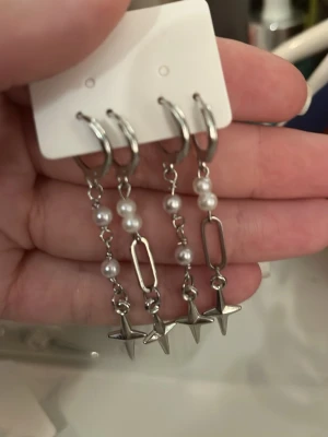 Silvriga örhängen med pärlor och stjärnor - Snygga silvriga örhängen med hängande kedjor, vita pärlor och stjärnformade hängen. Örhängena har även avlånga länkar som ger en cool och trendig look. Perfekta för att lyfta din stil med en unik touch.