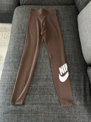 Bruna leggings från Nike - Snygga bruna leggings från Nike med hög midja och vitt Nike-logga tryckt på ena benet. Stretchigt material som sitter tajt och bekvämt. Perfekta för en sportig och avslappnad stil.