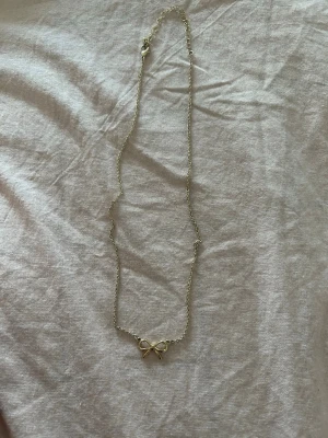 Guldigt halsband med rosett - Sött halsband i guldfärgad metall med en liten rosett som hänge. Kedjan är tunn och smidig, perfekt för att ge en gullig touch till din outfit. Passar dig som gillar enkla men fina detaljer.