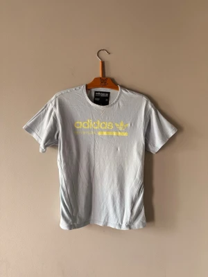 Ljusgrå Adidas t-shirt med gul logga - Snygg ljusgrå t-shirt från Adidas med gul tryckt logga och texten 'Built for Purpose' på bröstet. Klassisk rund hals och korta ärmar. Perfekt för en avslappnad och sportig stil.