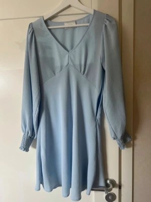 Ljusblå klänning från Neo Noir - Supersöt ljusblå klänning från Neo Noir med v-ringning och långa puffärmar med smock vid ärmsluten. Klänningen har en lätt utsvängd passform och är gjord i ett tunt, strukturerat tyg som ger en luftig känsla.