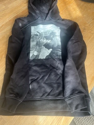 Svart hoodie med Tupac-tryck - Säljer en svart hoodie med stor svartvit Tupac-print på bröstet. Hoodien har huva med dragsko och en klassisk känguruficka framtill. Materialet ser ut att vara mjuk bomullsblandning, perfekt för en avslappnad streetwear-look.