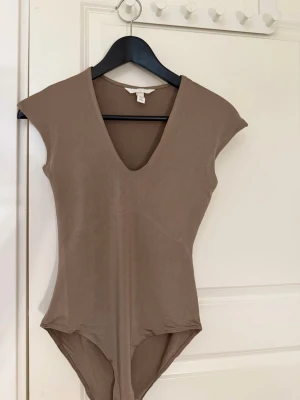 Brun/beige body med djup v-ringning från H&M - Säljer en stilren beige body med djup v-ringning och korta ärmar. Bodyn har en enkel och tight passform, bekväm känsla. Använts 2 gånger. 