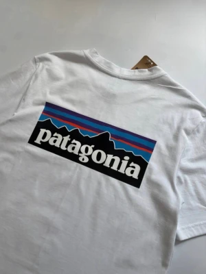 Vit Patagonia t-shirt med logga - Säljer en klassisk vit t-shirt från Patagonia med stor färgglad logga och bergmotiv på ryggen samt liten logga på bröstet. T-shirten har rund hals och korta ärmar, perfekt till vardags. Tillverkad i mjuk bomull för skön känsla.