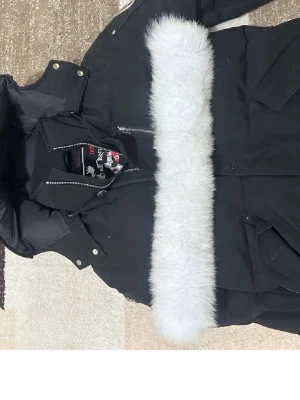 Svart dunjacka Moose Knuckles - Säljer en svart dunjacka från Moose Knuckles med stor vit fuskpälskrage på huvan. Jackan har dragkedja, tryckknappar och flera fickor. Perfekt för kalla vinterdagar och har en sportig, modern look.