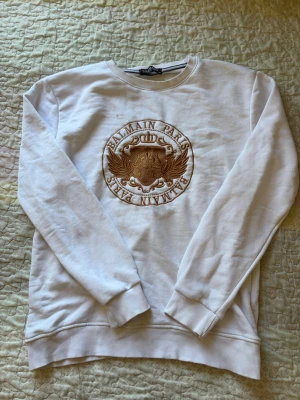Vit sweatshirt från Balmain Paris - Säljer en vit sweatshirt från Balmain Paris med stort broderat guldigt emblem på bröstet. Tröjan har rund halsringning, ribbade muddar och är tillverkad i mjukt material. Perfekt för dig som vill ha en clean men lyxig look.