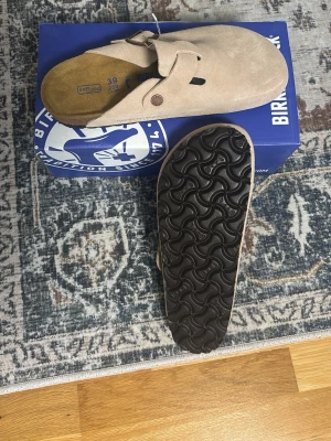Birkenstock Boston bs sand’ - Säljer ett par klassiska beige tofflor från Birkenstock i mocka med justerbart spänne och mjuk innersula. Yttersulan är i svart gummi med vågigt mönster för bra grepp. Perfekta för dig som vill ha både stil och komfort.