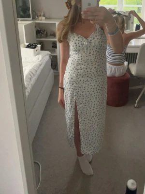 Söt långklänning i vitt med blått blommmönster  - Söt långklänning i vitt med blått blommönster från New Yorker. Klänningen har smala axelband med twistad detalj. Klänningen har en slits på ena sidan. På framsidan finns en liten liten defekt men syns knappt. 