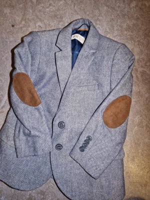 Barn 2-3 år blå Grå kavaj  med mockadetaljer H&M - 98 EUR, för barn 2-3 år 😊🩷. Stilren grå kavaj från H&M med klassiskt snitt och två knappar framtill. Kavajen har bruna mockalappar på armbågarna och mörkblått innerfoder. Materialet är vävt i diskret mönster och ger en dressad look. Perfekt för dig som vill ha en modern twist på klassikern.