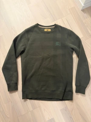 Olivgrön Burberry Tröja - Olivgrön Burberry Tröja med diskret rutmönster och broderad ryttare på bröstet. Tröjan har rund halsringning, ribbade muddar och är långärmad. Perfekt för en avslappnad och stilren look.