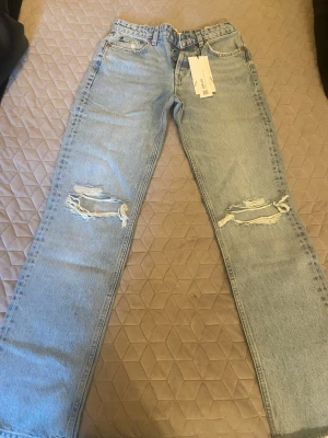 Zara Straight Mid rise full length jeans - Säljer ett par ljusblå jeans med raka ben och slitningar över knäna. De är helt oanvända. Prislappen sitter kvar. 