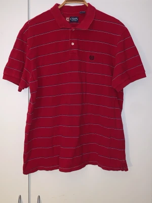 Röd pikétröja från Chaps XL - Snygg röd pikétröja från Chaps med tunna vita ränder och klassisk krage. Tröjan har två knappar vid halsen och är tillverkad i 100% bomull. Perfekt för en avslappnad och stilren look.
