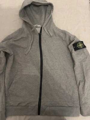 Grå zip hoodie från Stone Island - Snygg grå hoodie från Stone Island med dragkedja framtill, huva med dragsko och klassisk logopatch på vänster ärm. Tillverkad i mjukt sweatshirtmaterial och har två stora fickor framtill. Perfekt för en avslappnad och trendig look.