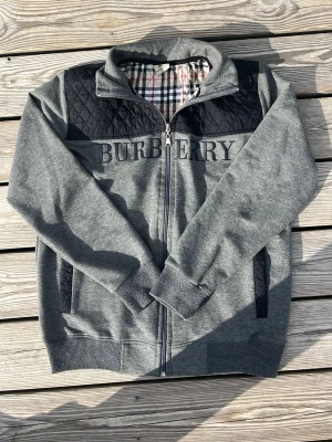 Grå ziptröja från Burberry - Snygg grå ziptröja från Burberry med quiltade svarta detaljer över axlarna och klassiskt rutigt foder. Tröjan har hög krage, ribbade muddar och två fickor framtill. Stor Burberry-logga broderad på bröstet. Perfekt för en stilren och avslappnad look.