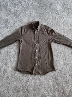 Brun skjorta från Morris - Snygg brun skjorta från Morris med klassisk krage och vita knappar framtill. Skjortan har lång ärm och en liten broderad logga på bröstet. Perfekt till jeans eller chinos för en clean look.