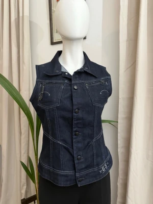 G-Star RAW Denim Mörkblå Jeansjacka - Snygg mörkblå ärmlös jeansjacka med markerade vita sömmar och två bröstfickor. Jackan har klassisk krage, knäppning framtill och detaljer som ger en cool, edgy look. Perfekt för att lyfta vilken outfit som helst.