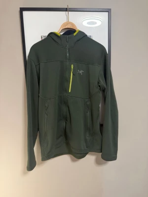 Grön hoodie från Arc'teryx L - Mörkgrön hoodie från Arc'teryx med huva och dragkedja framtill. Har en gulgrön bröstficka med dragkedja och två sidofickor. Tillverkad i mjukt fleece-material, perfekt för lager-på-lager och outdoor. Snygg detalj med logga på bröstet.