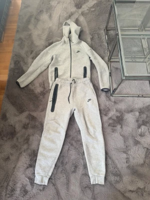 Grått Nike loungewear set med huva - Säljer ett grått loungewear set från Nike med matchande hoodie och byxor. Hoodien har dragkedja, två svarta dragkedjefickor och Nike-logga på bröstet. Byxorna har resår i midjan, snörning, svarta dragkedjefickor och Nike-logga på benet. Perfekt för chill dagar hemma.