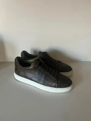 Mörkbruna sneakers med ormmönster -   Storlek: 40.5 — passar upp till 42 tack vare generös passform Färg: Mörkgrön/olivgrön med krokodilpräglad detalj vid hälkappan Material: Kalvskinn Sula: Vit gummisåla  Sneakers i äkta kalvskinn med en texturerad, reptilpräglad detalj vid hälkappan som ger dem ett unikt lyft. Mörkgrön ton som är sällsynt och lätt att kombinera med neutrala outfits — svarta byxor, beige chinos eller mörkt denim.  Skick: Bra skick. Normalt slitage på sulan, inga repor eller fläckar på ovandelen.  