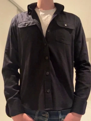 overshirt med bröstfickor - Stilren overshirt med knäppning framtill och två bröstfickor med lock och knapp. Skjortan har klassisk krage och raka ärmar. Perfekt att bära öppen över en t-shirt för en avslappnad look.            (Knappen på vänstra ärmen saknas)