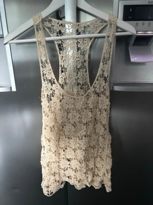 Beige spetslinne - Säljer ett superfint beige linne i spets med blommigt mönster. Linnet är ärmlöst och har en racerback-design som ger en cool och luftig känsla. Perfekt att styla över ett annat linne eller bikini för en trendig sommarlook.