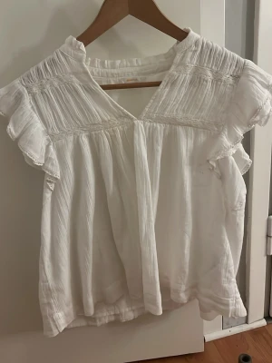 Fin blus ifrån Gina - Söt vit blus med v-ringning och korta volangärmar. Blusen har fina rynkade detaljer upptill och är gjord i ett lätt, luftigt tyg som känns skönt mot huden. Perfekt för en avslappnad och trendig look.