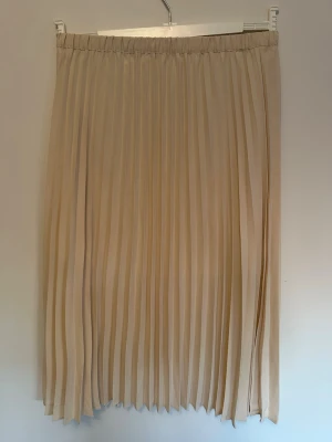 Plisserad beige långkjol (3/4 längd) - Snygg plisserad långkjol i 3/4 längd och som har en ljus beige/ cremefärgad nyans. Endast använd 1 gång. Kjolen har elastisk midja och ett luftigt, flowy tyg som ger en stilren och trendig look. Perfekt att matcha med en enkel topp eller stickad tröja.