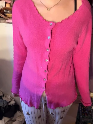 Rosa kofta med vågig kant - Säljer en söt rosa kofta med vågiga kanter och knappar framtill. Koftan har lång ärm och en feminin, figurnära passform. Perfekt att styla med jeans eller kjol för en trendig look.