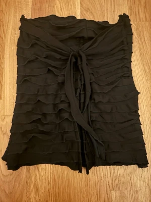 Svart omlott tubtopp med volang - Snygg svart tubtopp med omlottknytning framtill och volangiga vågade lager över hela toppen. Toppen är ärmlös och har en trendig, croppad känsla. Perfekt för dig som vill sticka ut med en cool och stilren look. Om du intreserad jag vill bara förvarana att dom här byxorna är från en liten företag som hetter jennifer.