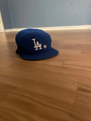 Blå LA keps med broderad logga - Snygg blå keps från New Era med LA-logga broderad i vitt framtill och ett litet hjärta bredvid. Klassisk flat peak och broderade ventilationshål. MLB-logga och New Era-märke på sidan. Perfekt för dig som gillar streetwear och sportig stil. Riktigt bekväm att ha på sig!