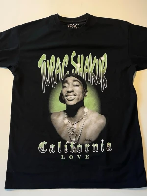 Pull & Bear Tupac Shakur t-shirt - Svart t-shirt med tryck av Tupac Shakur. Trycket inkluderar texten "Tupac Shakur California Love" i gröna och vita nyanser. Tröjan har korta ärmar och en rund halsringning.  Modellen är slutsåld i butik och går inte längre att få tag på!