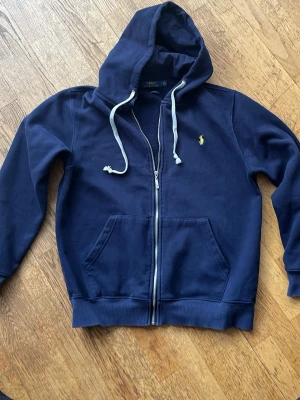 RL Mörkblå zip hoodie med huva - Mörkblå hoodie med dragkedja från Polo Ralph Lauren. Klassisk modell med huva och vita dragsnören, två fickor framtill och gul broderad logga på bröstet. Tillverkad i mjukt bomullsmaterial, perfekt för en avslappnad stil.
