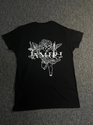 Svart AMIRI t-shirt med ängeltryck - Snygg svart t-shirt från AMIRI med stort vitt ängeltryck på ryggen och coolt AMIRI-tryck med färgstänk på bröstet. Klassisk passform och korta ärmar. Perfekt för dig som vill sticka ut med en unik design.