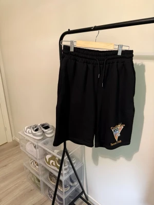 Svarta shorts från Casablanca - Svarta shorts från Casablanca med trycket 'Tennis Club' och en färgglad grafisk detalj på ena benet. Shortsen har elastisk midja med snörning och är tillverkade i mjuk bomull, perfekta för en avslappnad och sportig stil.