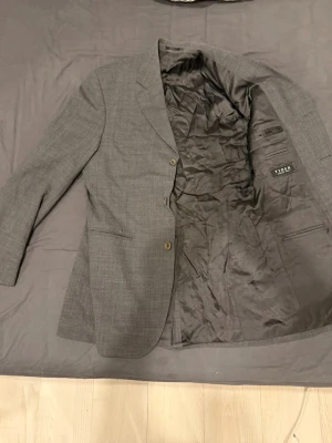 Grå rutiga kavajer herr - Två stilrena grå kavajer med diskret rutmönster och klassisk passform. Båda har två knappar framtill, fickor och helsvart foder. Perfekta för en dressad look och passar till skjorta eller t-shirt. Märket Tiger syns i ena kavajen. Storlek 46 och 50