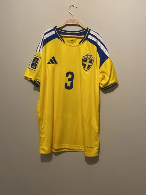Svensk landslagströja Lindelöf #3 - Gul svensk landslagströja från Adidas med blå detaljer, nummer 3 och namnet Lindelöf på ryggen. Tröjan har FIFA-märke på ärmen, klubbmärke på bröstet och klassiska Adidas-ränder på axlarna. Perfekt för fotbollsfans som vill stötta Sverige. 