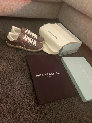 Bruna Philippe Model sneakers - Snygga bruna Philippe Model sneakers med vita detaljer och vita skosnören. Skorna har en låg profil och beige sula med märkets logga på sidan och under sulan. Tillkommer originalkartong och dustbag. Perfekta för dig som gillar stilrena och trendiga sneakers.