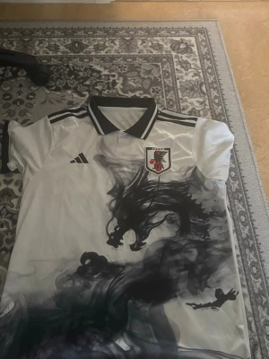 Japan fotbollströja Adidas svartvit - Säljer en snygg Japan fotbollströja från Adidas i vitt med svart rök-mönster och svarta detaljer. Tröjan har krage, korta ärmar och Japan-emblem på bröstet. Perfekt för dig som gillar unika sportkläder och vill sticka ut på planen.