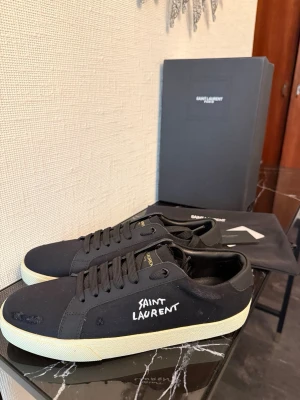 Svarta Saint Laurent canvas sneakers - Svarta sneakers från Saint Laurent i canvas med vit logga broderad på sidan och vita sulor. Klassisk låg modell med svarta skosnören och diskreta guldfärgade detaljer på plösen. Perfekta för en stilren och trendig look.