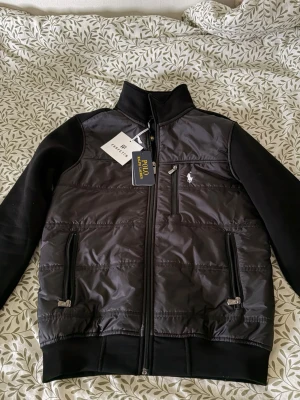 Svart cardigan från Polo Ralph Lauren - Snygg svart cardigan från Polo Ralph Lauren med dragkedja framtill, tre fickor med dragkedja och diskret logga på bröstet. Jackan har en mix av material med glansig front och mjuka ärmar. Perfekt för dig som vill ha en stilren och sportig look.