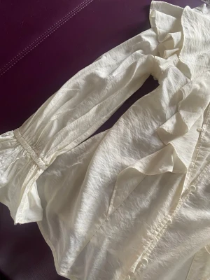 Vit volangblus från Indiska - Stilren vit blus från Indiska med långa ärmar och volangdetaljer längs knäppningen och axlarna. Blusen har knappar framtill, använd 1 gång. Jag vill sälja den eftersom den passar inte min still längre 💗
