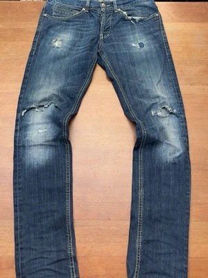 Dondup jeans - Säljer ett par blå jeans med slitningar och hål på knäna samt snygga slitna detaljer framtill och baktill. Jeansen har klassisk femficksmodell, kontrastsömmar och raka ben. Perfekta för en avslappnad och trendig look.