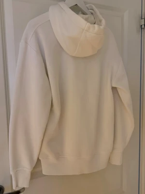 Vit oversized hoodie med ficka - Säljer en clean vit hoodie med stor känguruficka framtill och dragsko i huvan. Hoodien har ribbade muddar vid ärmar och nederkant, samt en avslappnad passform. Perfekt för en chill och stilren look. Materialet känns mjukt och bekvämt.