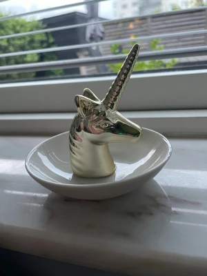 Unicorn ringhållare i keramik - Snygg ringhållare i form av ett enhörningshuvud med horn i guld och fat i vit keramik. Perfekt för att förvara ringar eller små smycken på ett stilrent och lekfullt sätt. Enhörningen har en blank finish och fatet är runt.