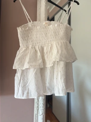 Vit volangig linnetopp med tunna band - Supersöt vit linnetopp med tunna axelband och volangdetaljer. Toppen har smockad överdel och ett skirt, lätt material med diskret glittrigt mönster. Perfekt för varma sommardagar och ger en romantisk vibe. Skriv innan köp