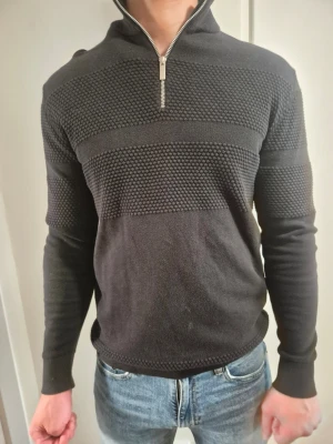 Mörkgrå stickad tröja med halv dragkedja - Snygg mörkgrå stickad tröja med halv dragkedja och strukturstickade partier över bröst och armar. Tröjan har hög krage och långa ärmar, perfekt för lager på lager. Passar till jeans för en avslappnad look.