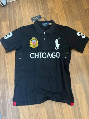 Polo ralph lauren - chicago black - Svart pikétröja från Polo Ralph Lauren med vit logga och texten CHICAGO framtill. Broderad patch i gult och rött på bröstet samt siffran 3 på ärmar och rygg. Klassisk krage och knappar framtill. Perfekt för en sportig och stilren look.