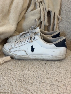 Vita Polo Ralph Lauren sneakers - Klassiska vita sneakers från Polo Ralph Lauren med mörkblå detaljer och logga på sidan. Skorna har lågt skaft, vita snören och är tillverkade i slätt läder med textilfoder. Perfekta för en clean och stilren look. De är ganska använda och har något litet hål i insidan som du kan se på sista bilden☺️